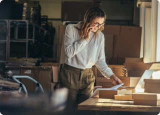 Woman wearing glasses and business clothing standing over desk, talking on the phone, looking at a piece of paper